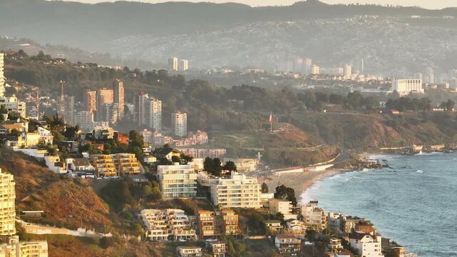 Drone telephoto compressed zoom shot with parallax effect moving sideways over cliffs showing Renaca beach and Vina del Mar skyline at sunset in Chile