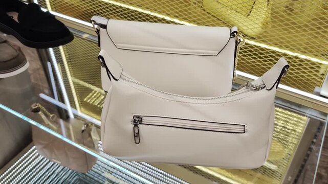 Two white, modern design handbags showcased on clear glass shelf, featuring silver zippers and hardware, one bag with visible rear pocket, second bag with flap closure, upscale shopping environment