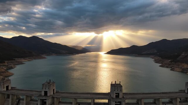 Sun rays breaking through clouds over dam reservoir with mountains on horizon and reflective water surface creating epic lighting effect in dramatic natural landscape sky reflection