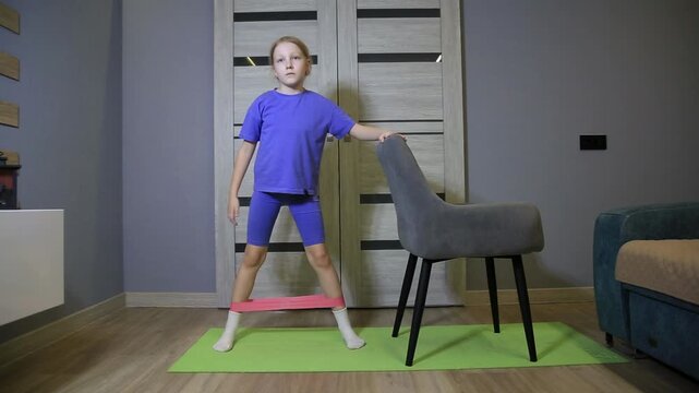 Caucasian child with blonde hair performs leg spread with resistance band. Exercise routine at home for muscle development and healthy lifestyle.
