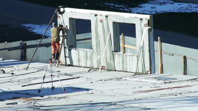 Construction worker standing on ladder next to large prefabricated concrete wall panel with two window openings. Worker engaged in installation of building frame section on rooftop. Outdoor scene duri