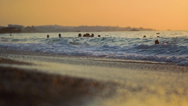 Low angle view of ocean waves breaking on beach shore at golden hour. Sun reflection on water surface and wet sand. People enjoy swimming in water with building outline on horizon. Warm colors of even