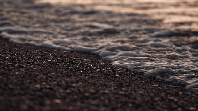 Ocean waves gently wash dark pebble beach. Foamy water recedes, exposing wet stones. Warm light on water highlights rock texture. Calm coastal scene, natural beauty. Slow motion