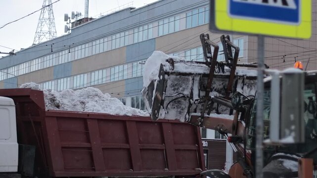 Heavy machinery clearing city streets of snow after winter storm. Front end loader bucket lifts white snow, dumping it into large red dump truck. Urban environment, essential service activity daylight