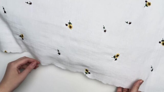 Hands display white lightweight cotton muslin textile decorated with small sunflower embroidery showcasing delicate weave breathable structure soft surface and fine material details suitable for summe