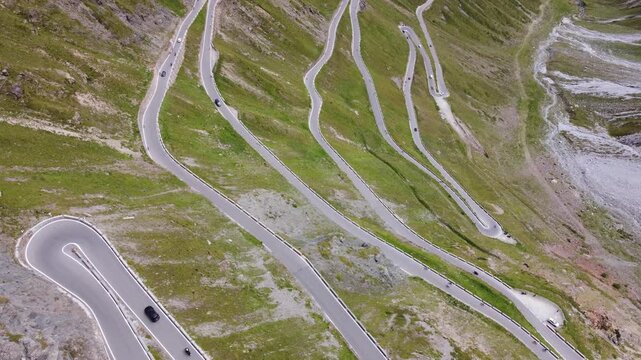 4K aerial drone view from above of the iconic Stelvio Pass road in the Italian Alps. Sharp serpentine curves and hairpin turns carved into the steep green mountain slope.