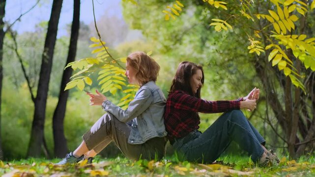 Two young women, a blonde and a brunette, are sitting back to back on a lawn among autumnal trees and using their smartphones. A loving LGBT couple enjoys a sunny autumn day in the park.