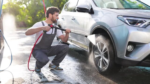 A caucasian man in overalls crouches to pressure wash a silver car, creating a lot of foam and spray on the ground