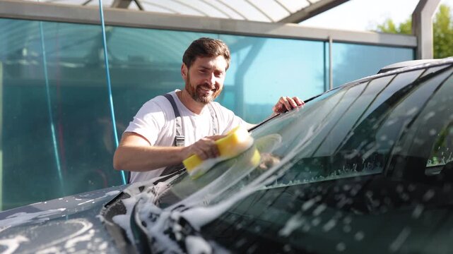 A smiling man in overalls is actively washing the windshield of a car with a yellow sponge and soap suds
