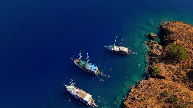 Aerial view of traditional gulet boats anchored near a rocky coastline in clear turquoise water, ideal for summer travel, coastal tourism, sailing, vacation and Mediterranean lifestyle content.