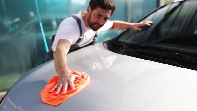 A smiling Caucasian man in overalls is meticulously polishing the hood of a car with an orange microfiber cloth, ensuring a spotless finish