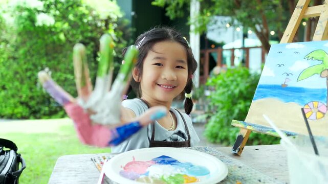 Cute happy Asian little girl showing her colorful painted hand, smiling child enjoying creative art workshop and beach canvas painting activity in the park