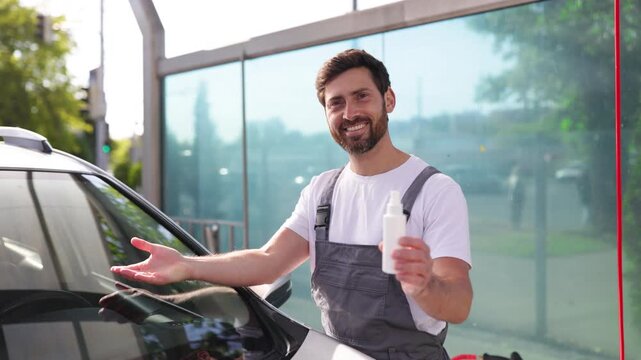 A smiling caucasian man in work overalls holds a spray bottle and gestures towards a car windshield, ready for cleaning
