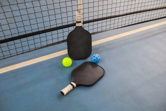 Two Pickleball Paddles And Two Wiffle Balls Rest On An Indoor Court Surface