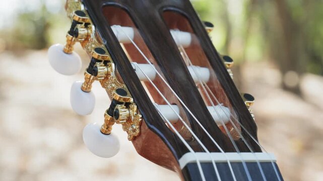 Close-up of the guitar head of a classical guitar with gold pegs. Part of a classical guitar on a blurry background in the park. Head of guitar with nylon strings. Slow motion 4k footage.