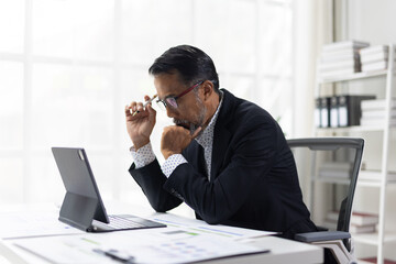 Naklejka premium Asian businessman concentrating and thinking while reviewing financial documents and market data on a tablet device at his office desk, problem-solving and strategy.
