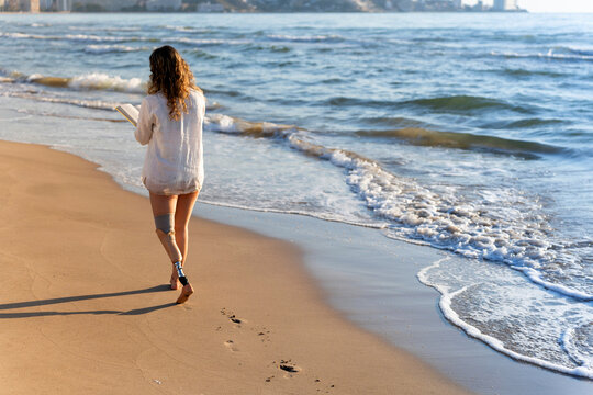 Woman with prosthetic leg walking on valencia beach reading a book