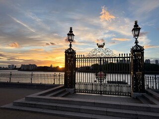 Fototapeta premium Ornate Riverside Gate: Intricate black wrought iron gate with vintage lanterns standing in silhouette against a beautiful glowing sunset by the river