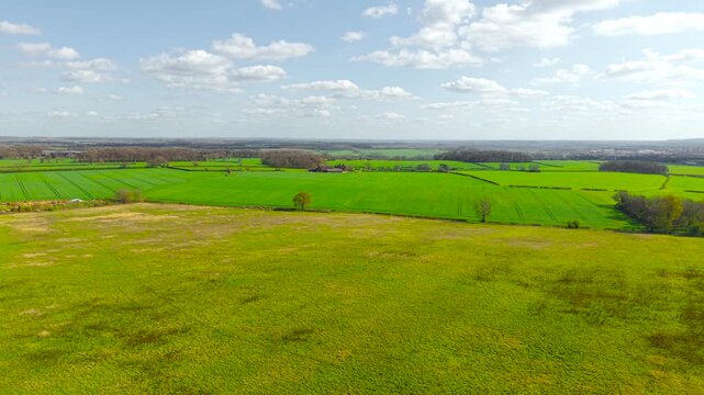 Open grassy field in foreground transitioning into structured agricultural plots with scattered trees and distant farms. Elevated view emphasizing texture of land seasonal growth and rural simplicity