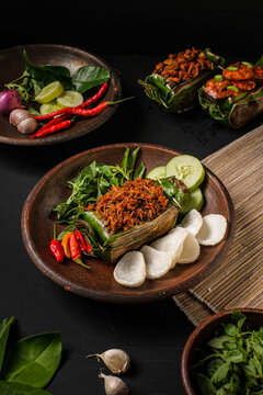 Authentic Nasi Bakar Teri, traditional Indonesian grilled rice wrapped in banana leaf with spicy savory anchovies. Plated on rustic clay dish with crackers and fresh cucumber.