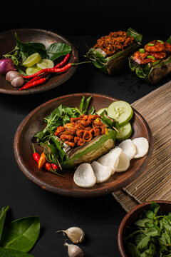 Delicious Nasi Bakar Usus, Indonesian traditional grilled rice wrapped in banana leaf topped with spicy chicken intestines. Plated on a rustic clay dish with crackers and basil.