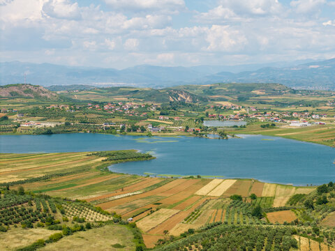 Aerial view of a blue lake surrounded by a patchwork of agricultural fields, olive groves, and small rural villages under a cloudy sky in Albania.