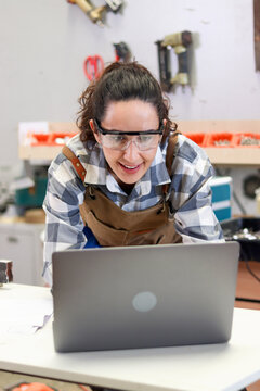 Happy smiling carpenter woman wearing safety glasses goggles and apron, working on her laptop computer. A beautiful female craft worker creating wooden furniture in a woodworking carpentry workshop.