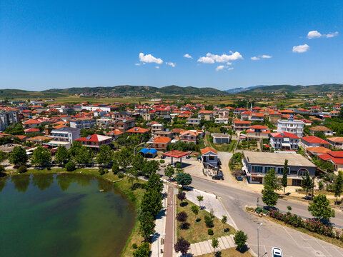 Aerial view of the town of Gradishte featuring red-roofed houses, a small lake, and lush green trees under a clear blue sky in Gradishte, Fier County, Albania.