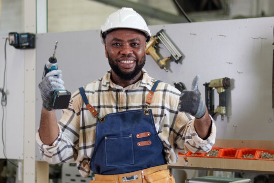Portrait of happy African carpenter wearing white helmet and apron, holding an electric drill and giving thump up. Craftsman worker man creating wooden furniture in woodworking carpentry workshop.