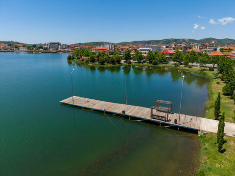 Aerial view of a wooden pier on the calm waters of a lake with the town buildings and green trees under a clear blue sky in Gradishte, Fier County, Albania.