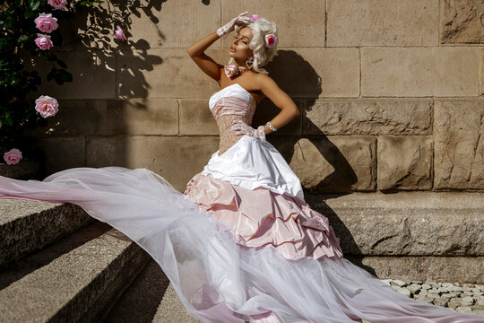 Ethereal Rococo Inspired Fashion Portrait: Elegant Woman in Luxurious Pink and White Ball Gown Posing Against Ancient Stone Wall with Roses