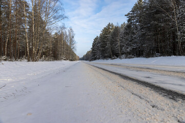 Obraz premium a narrow track covered with snow from cars in the forest in the winter season, different types of trees grow on the side of the road against the blue sky