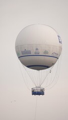 Biały, okrągły balon widokowy na uwięzi unosi się wysoko na tle zamglonego nieba. Pod wielką powłoką ozdobioną grafikami widać wiszącą gondolę dla pasażerów oraz stabilizującą linę. © tomo81