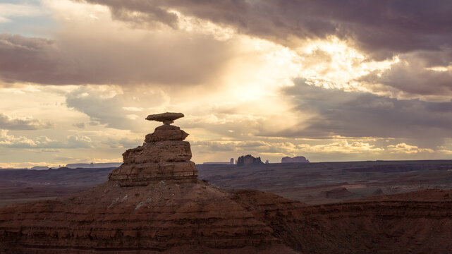 Aerial view of the Mexican Hat rock formation under a dramatic sky with sunbeams breaking through clouds over the desert landscape in Mexican Hat, Utah, United States.
