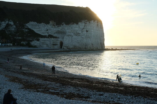Aerial view of the towering white chalk cliffs and pebble beach with people walking along the shore and swimming at sunset in Yport, Normandy, France.