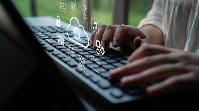 Close-up image of hands typing on a laptop keyboard with cloud icons floating above, representing digital technology and remote work in a modern workspace. Gantry