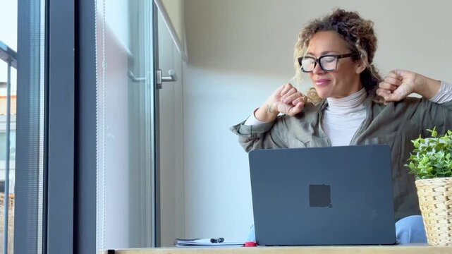 Happy adult woman finishing her work on a laptop at home, smiling and stretching her arms in relief and satisfaction while sitting at a desk near a window with a nice bright modern light