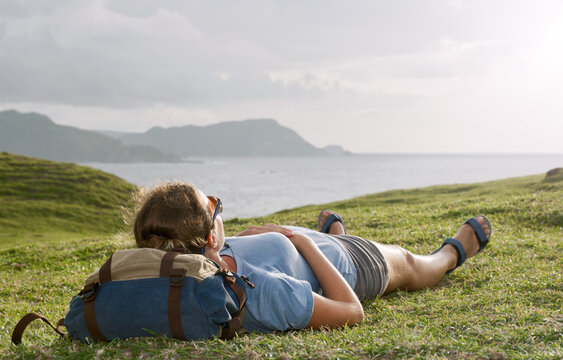 Woman with backpack lying on green grass hill, looking at sea and mountains. Lifestyle, outdoor adventure and travel concept.