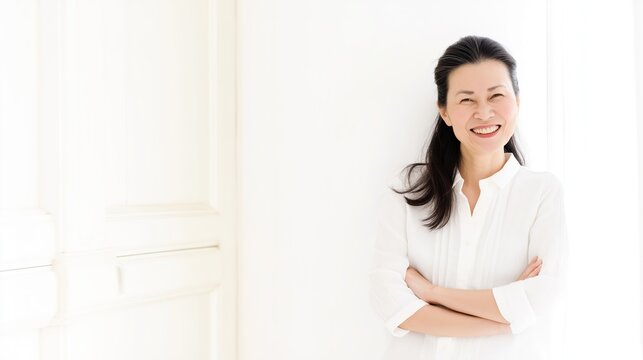 Lifestyle portrait of vibrant beautiful Japanese woman in her 50s, confident smiling pose with crossed hands, elegant casual outfit, bright airy white room, natural and studio soft lighting, clean col