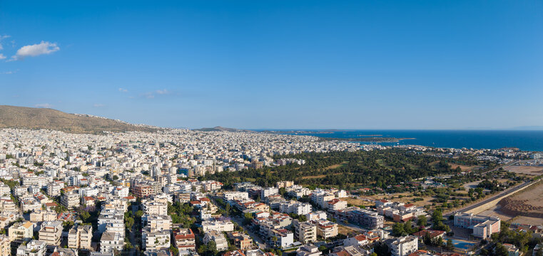 Aerial view of the dense urban landscape featuring white residential buildings, a large green park, and the blue Saronic Gulf under a clear sky in Glyfada, Athens, Greece.