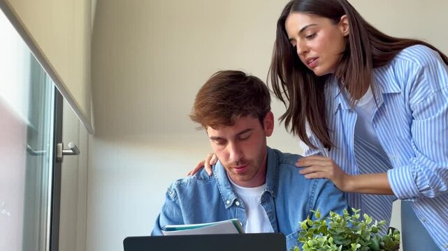 Caring young girlfriend supporting her stressed boyfriend while he is working on his laptop from home, helping him with some notes and giving him some much needed emotional support