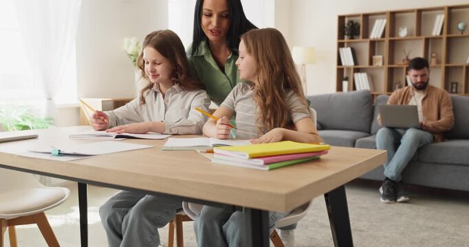 Family children studying homework with parents mother father. Daughters write as mother guides while father works on a laptop, showing supportive home education. Simple family study idea now.