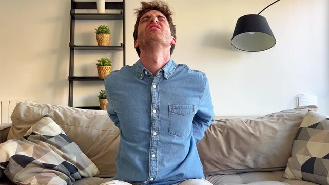 Frustrated young man sitting on a sofa in the living room feeling a strong backache and massaging his sore muscles, showing facial expressions of discomfort and suffering from the injury
