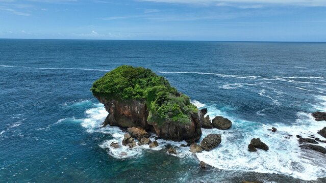 Aerial view of a lush green rocky islet surrounded by crashing white waves in the blue ocean at Jungwok Beach Girisubo, Special Region of Yogyakarta, Indonesia.