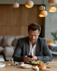 Man sits at table, focused on phone. Warm lights hang above, soft and ambient. Food rests beside him, untouched. He seems thoughtful, perhaps distracted. Ideal for scenes of modern disconnect © lashkhidzetim