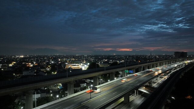 Aerial view of a multi-level highway overpass with car light trails at dusk under a dark cloudy sky in Bekasi Barat, West Java, Indonesia.