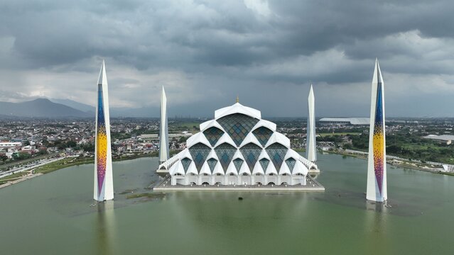 Aerial view of Masjid Raya Al Jabbar featuring its modern tiered architecture and four tall minarets surrounded by water under an overcast sky in Kecamatan Gedebage, Jawa Barat, Indonesia.