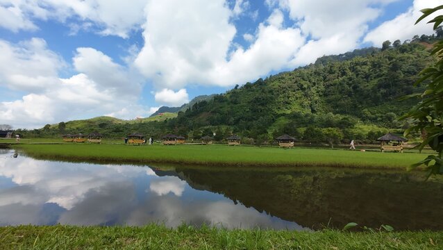 Aerial view of Situ Rawa Gede lake with wooden gazebos and people on the grassy shore reflected in calm water with lush green hills in Sukamakmur, West Java, Indonesia.
