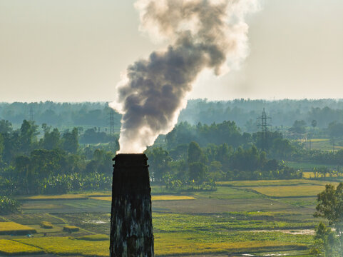 Aerial view of a tall industrial chimney emitting thick white smoke over lush agricultural fields and rural landscape in Bogura, Rajshahi Division, Bangladesh.