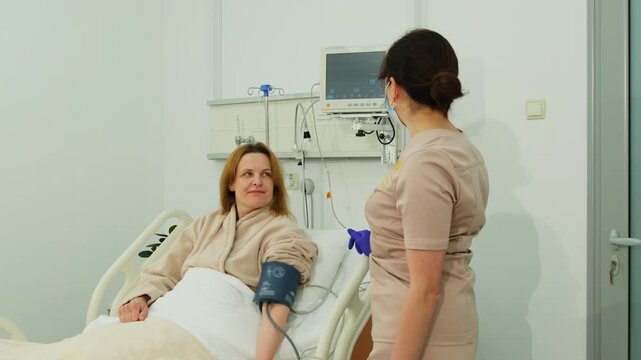 Woman patient looks at nurse from hospital bed during pressure test. Nurse observes cuff result following woman response near white bed